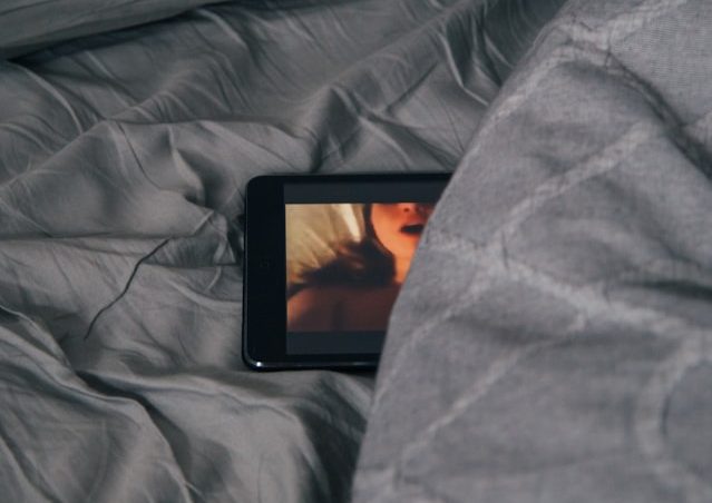 une tablette avec une image d'un visage de femme blanche bouche ouverte. La tablette est posée dans un lit aux draps gris, à moitié couverte par la couette