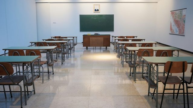 photo d'une salle de classe vide des rangées de bureaux et de chaises à gauche et à droite. au fond, un bureau d'enseignant·es et un tableau noir. Au dessus du tableau, un portrait (dont on ne voit pas le visage) sur le mur de droite, une carte géographique. by Ivan Aleksic